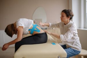 A physiotherapist applies kinesiology tape to a young woman's back during a therapy session.